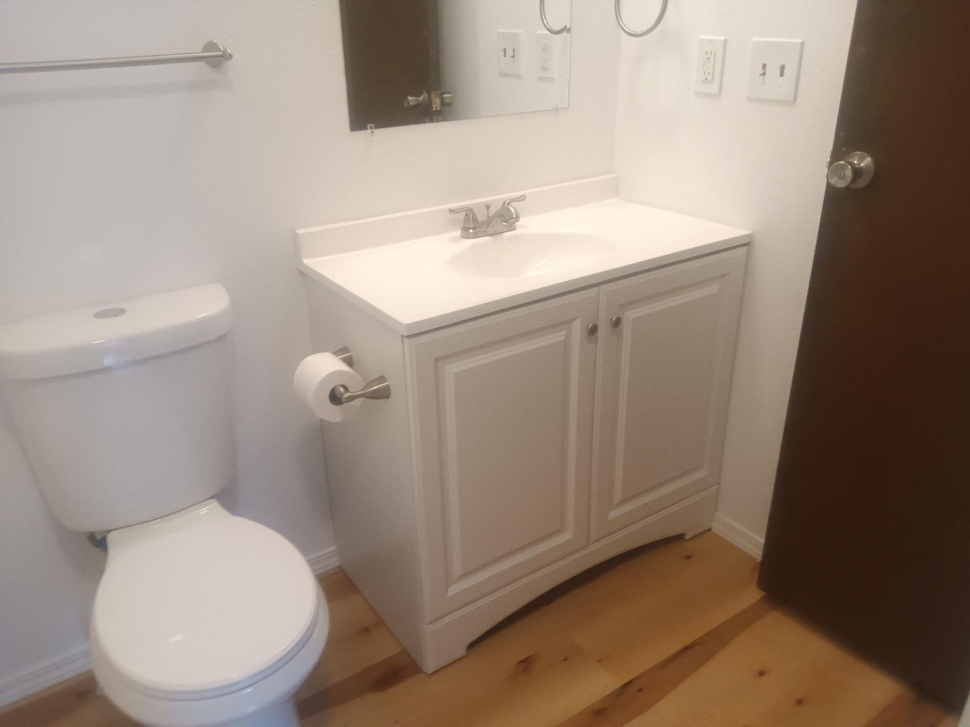 Small bathroom with toilet, white vanity sink, mirror, and wooden floor beside a dark wood door
