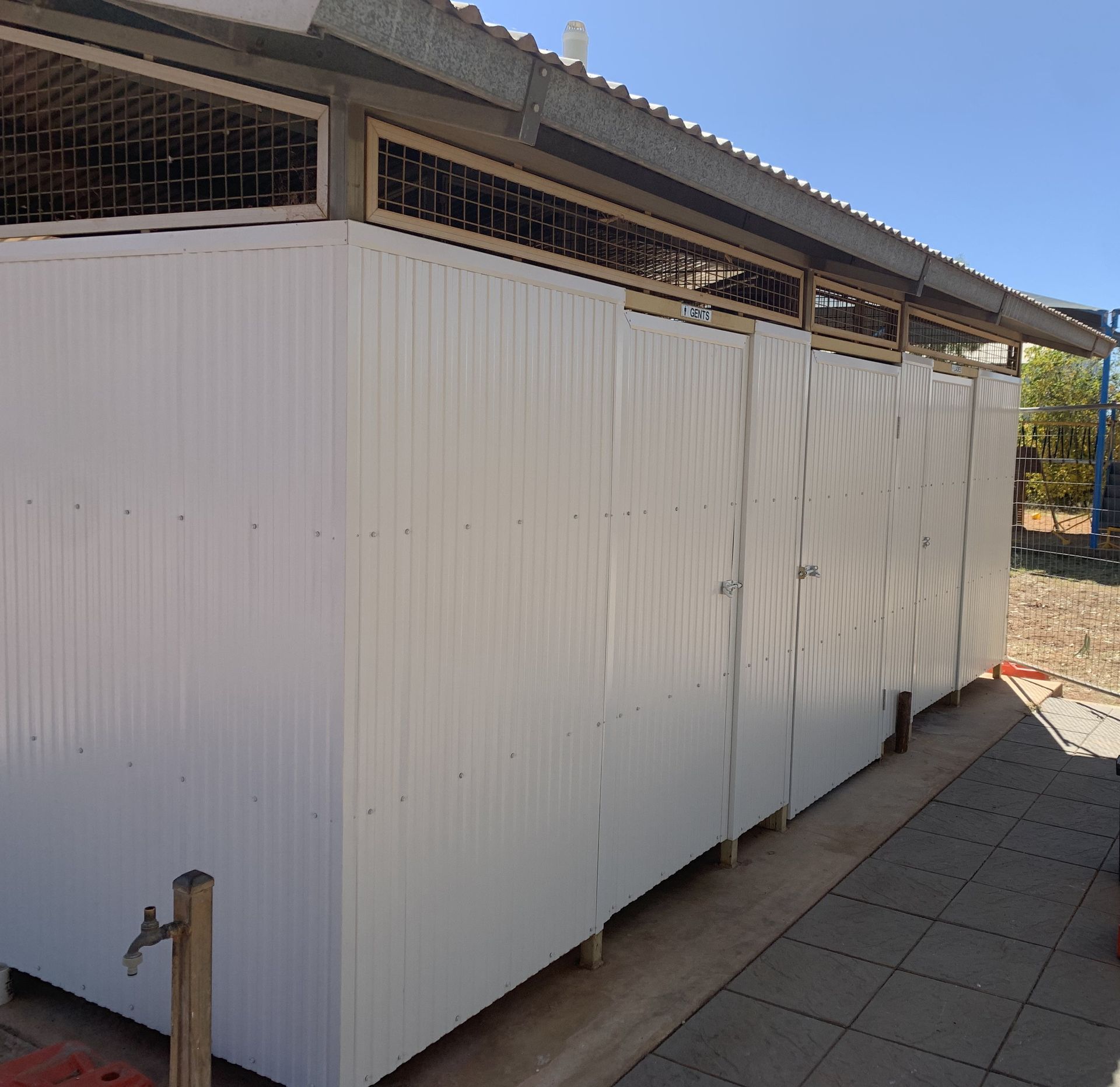 A row of white, corrugated metal outdoor toilet stalls with a mesh ventilation strip under a slanted metal roof.