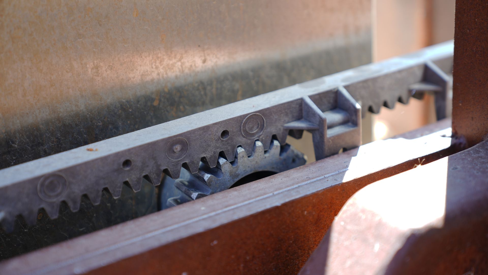 Close-up of a metal gear rack and toothed mechanism inside a machine, lit by sunlight.
