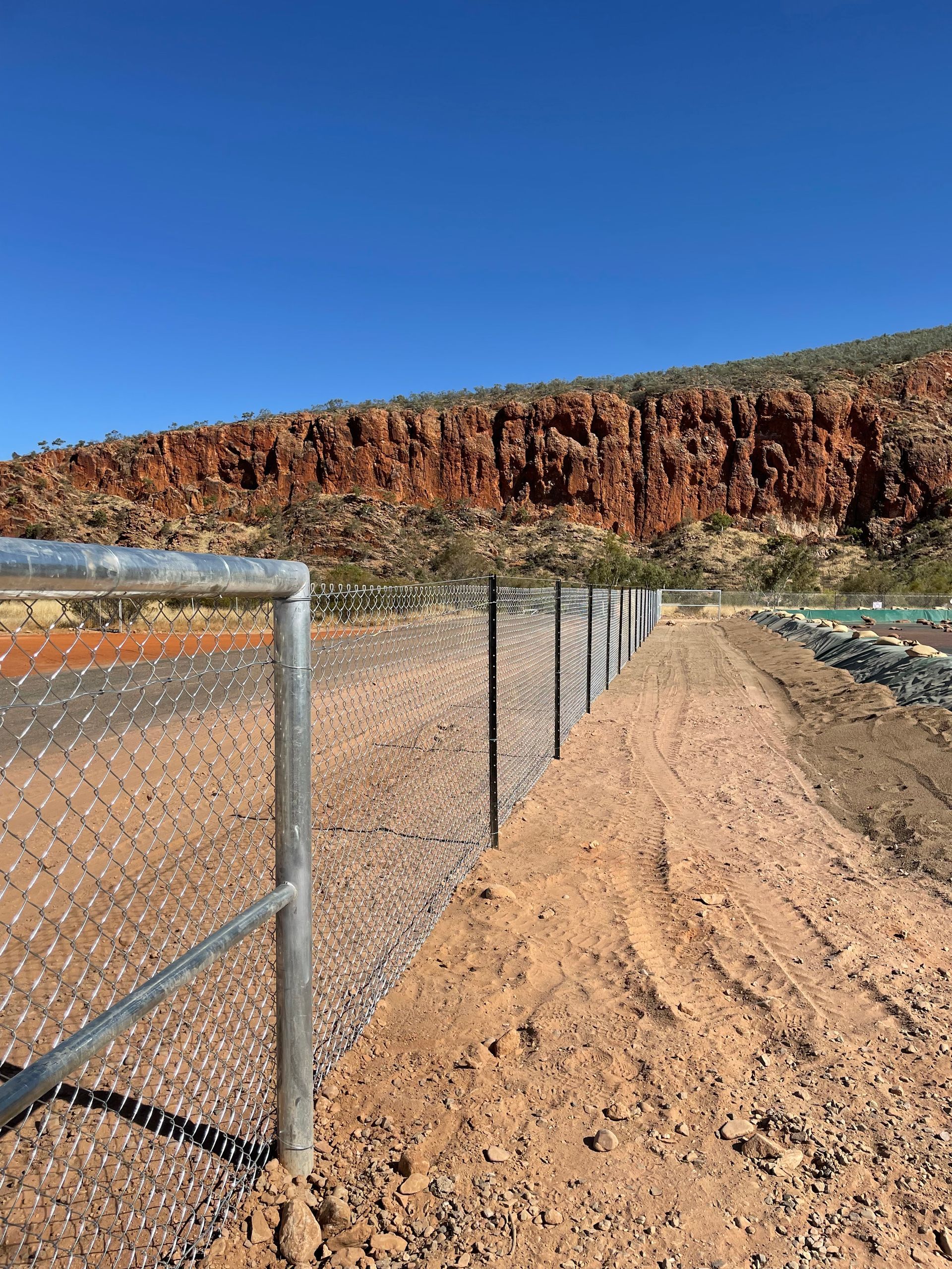Chain-link fence beside a dirt path under a blue sky, with a rocky red cliff in the background
