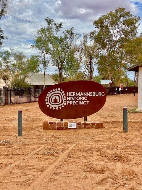 A brown, oval sign reads "Hermannsburg Historic Precinct" in a dusty, outdoor setting with trees under a cloudy sky.