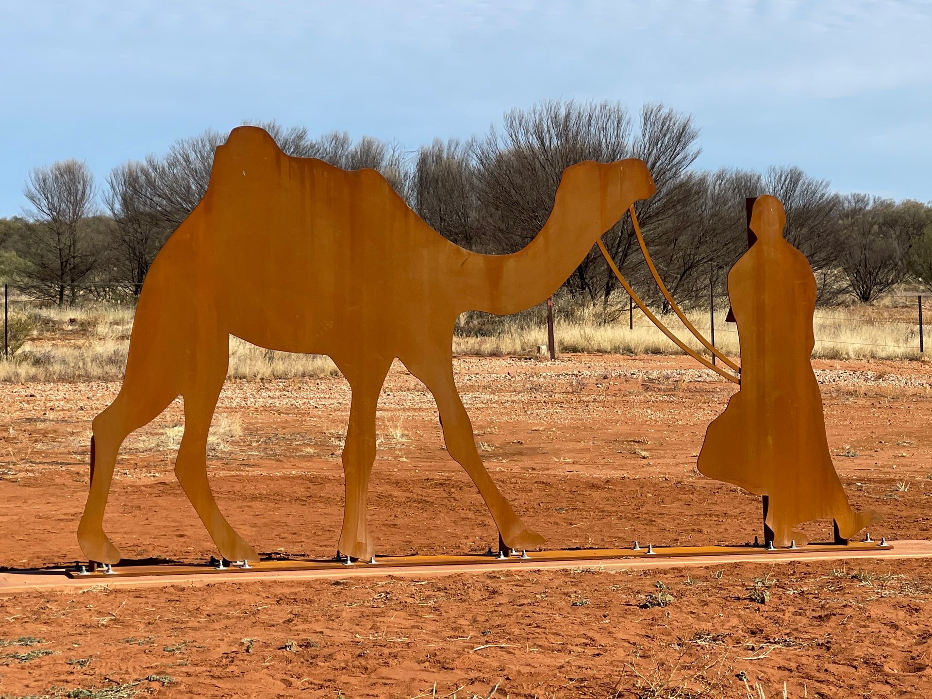 A rusted metal silhouette of a camel and a person leading it stand on red dirt against a backdrop of trees and blue sky.