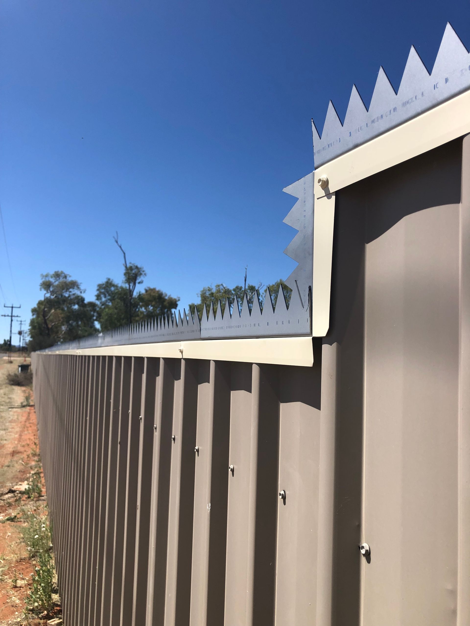 Brown fence with pointed top along a sunny exterior wall under a clear blue sky