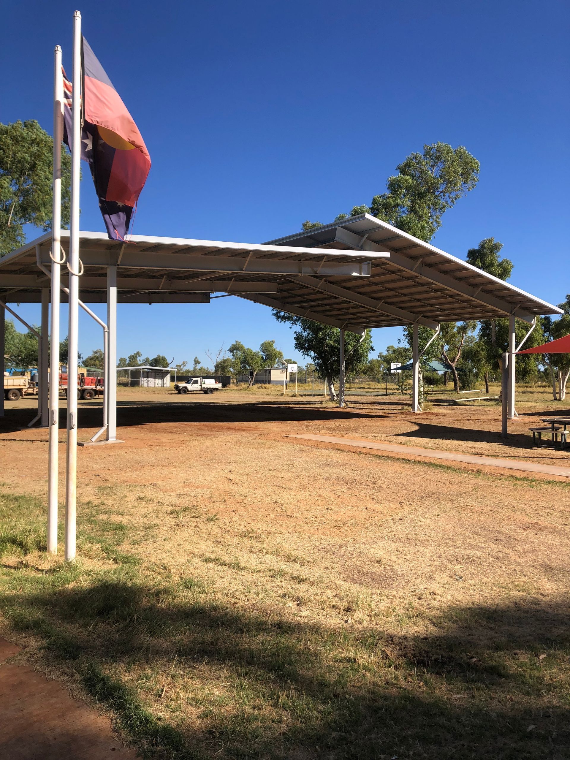 A solar panel shelter structure standing over a dirt lot next to a flagpole with a flying flag under a bright blue sky.
