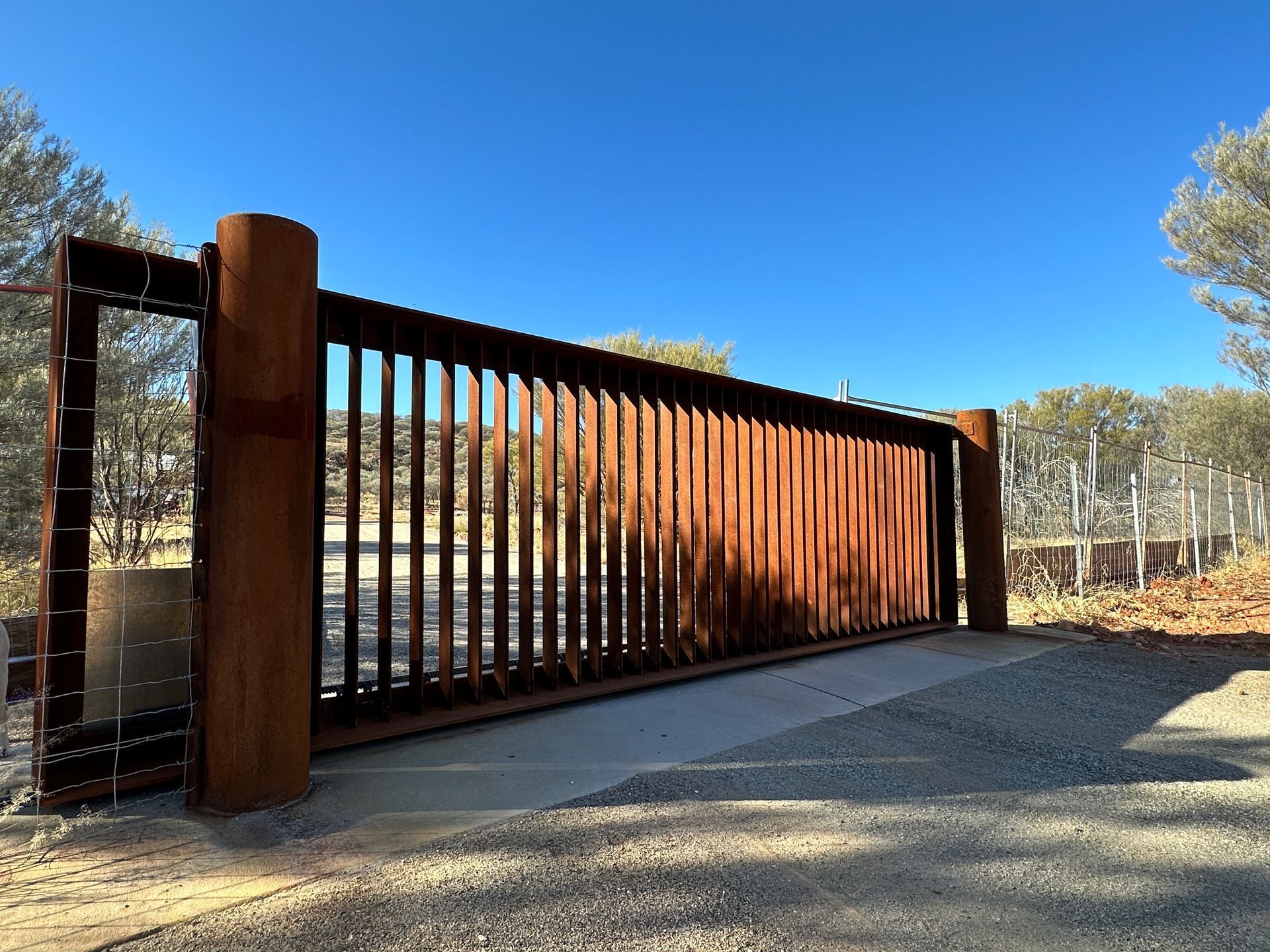 Rust-colored metal sliding gate beside a gravel driveway under a clear blue sky