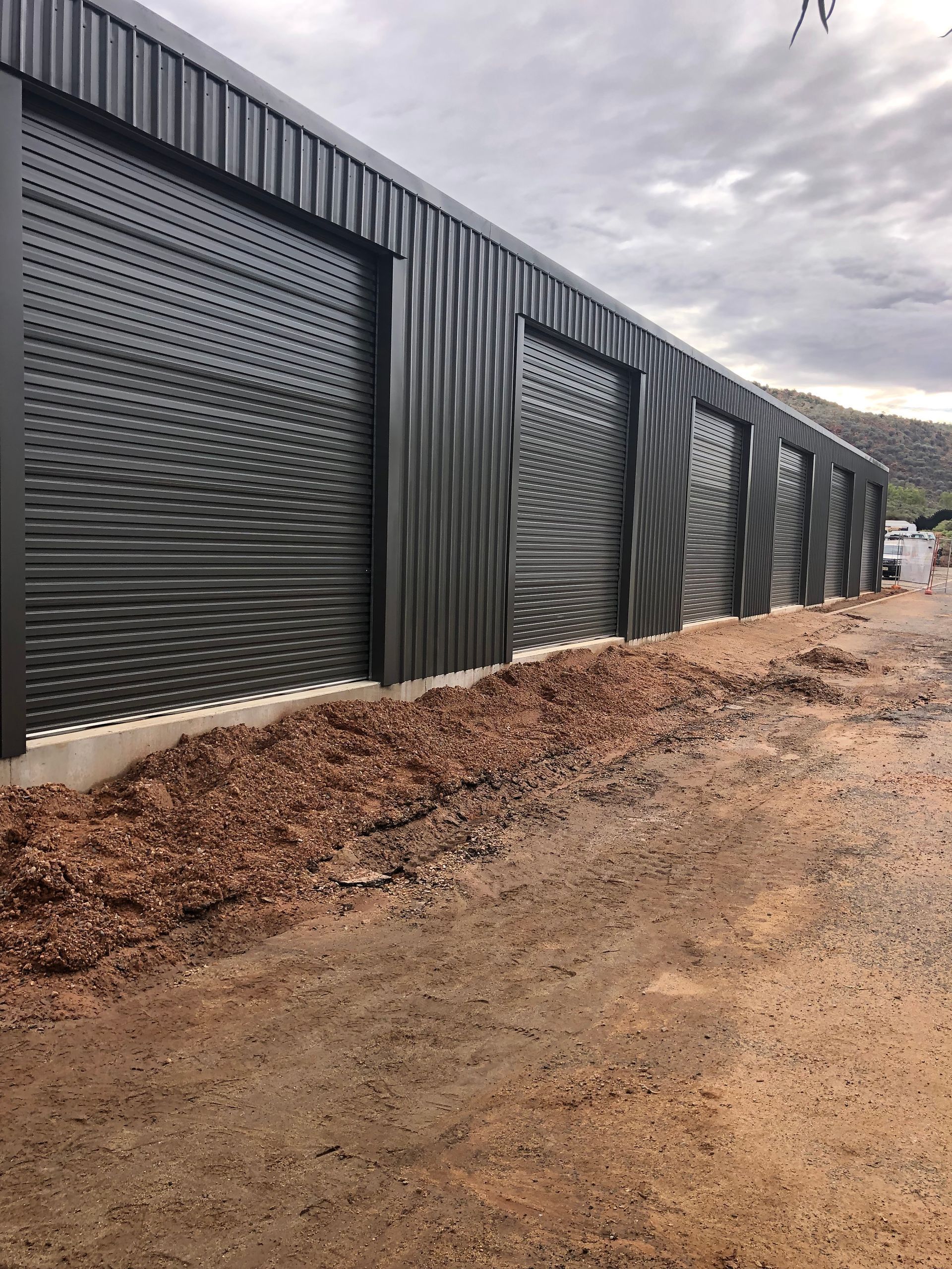 A modern black metal storage building with multiple roll-up doors, set against a cloudy sky and dirt ground.