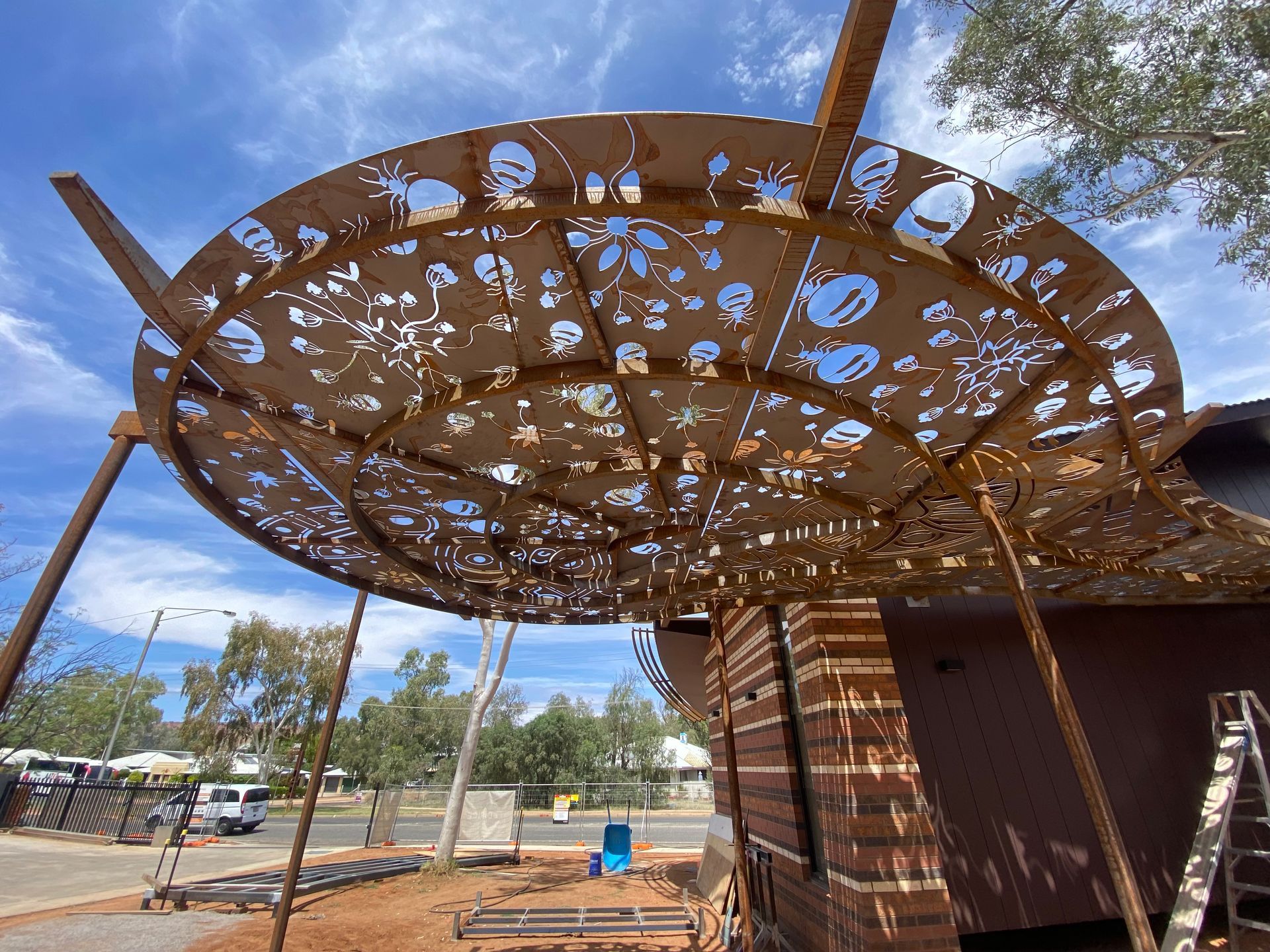 A rusted metal canopy with intricate, laser-cut floral patterns shades the side of a building under a blue sky.