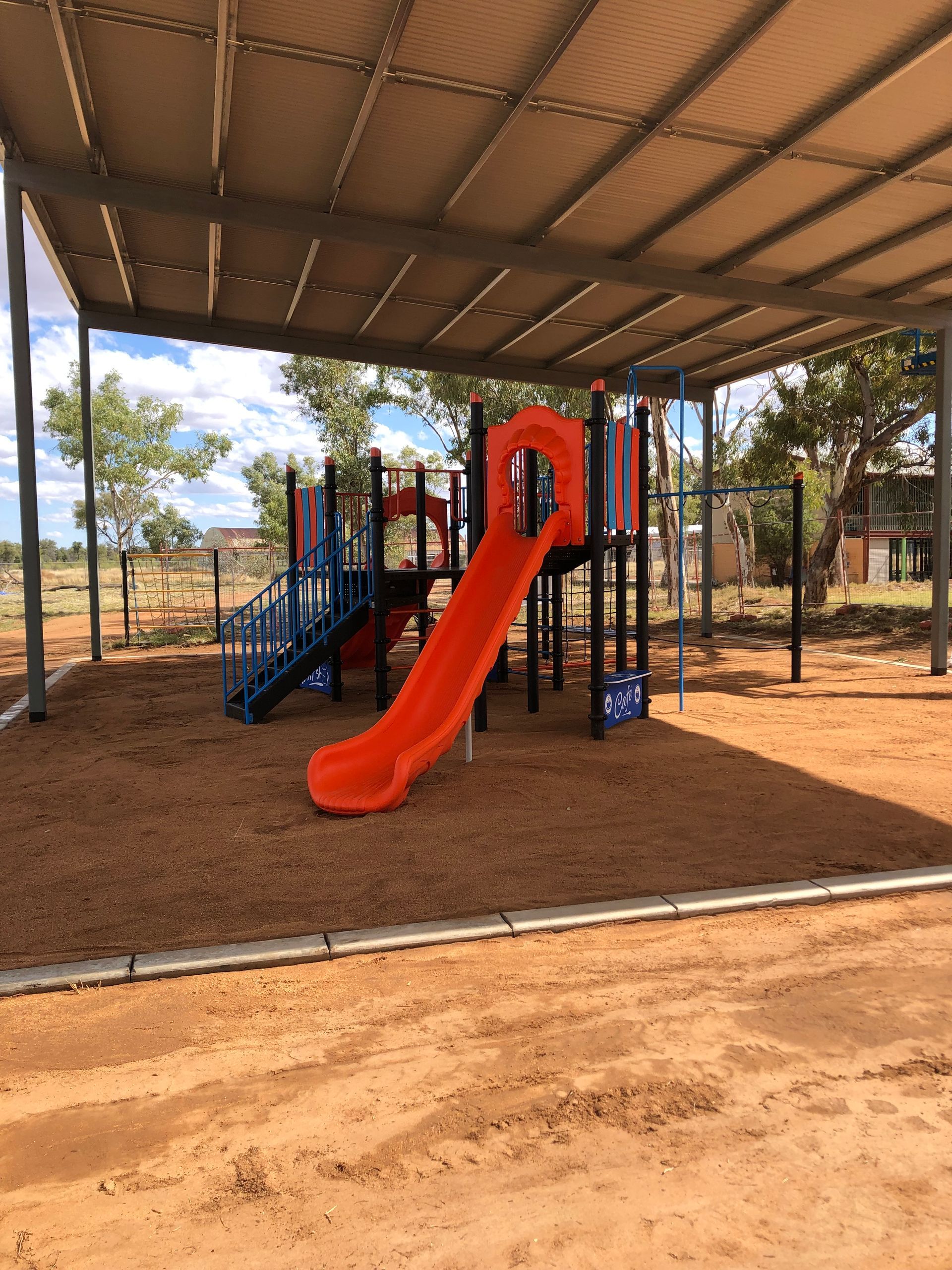 A playground with a bright orange slide and blue steps under a metal pavilion on a dirt ground surface.