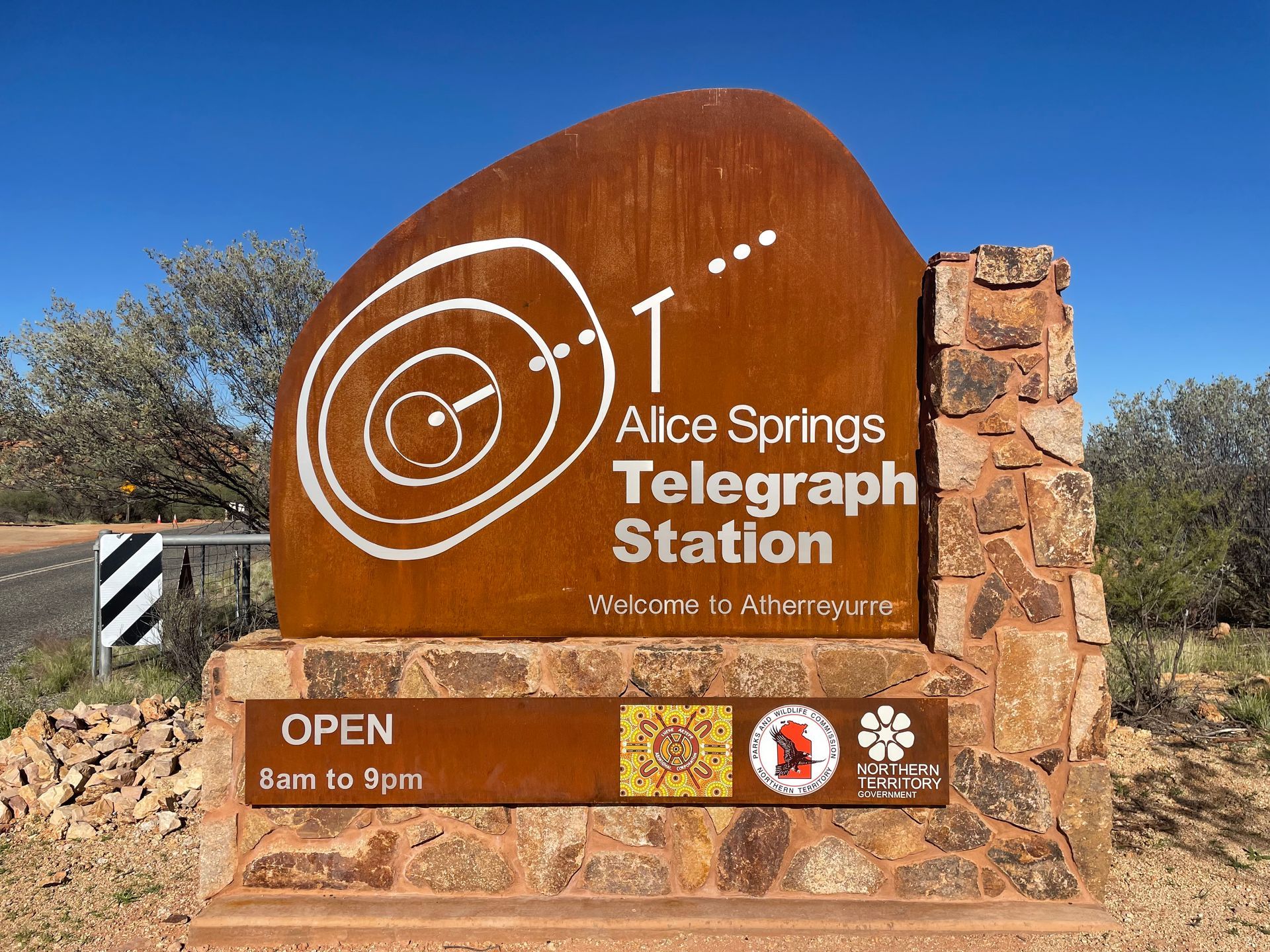 A rusted metal sign for the Alice Springs Telegraph Station set in stone, noting it is open from 8am to 5pm.