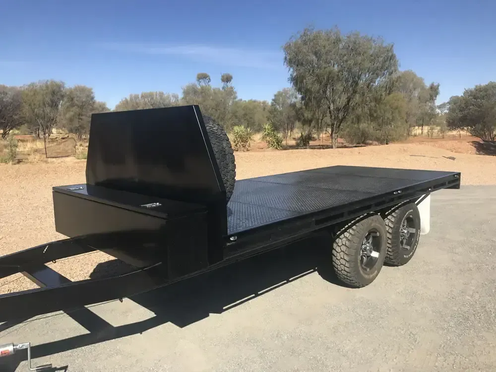 Black flatbed trailer with front box and tandem wheels parked on a road in a dry, wooded area