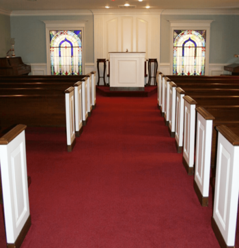 Empty church sanctuary with red carpet aisle, wooden pews, and stained-glass windows around the pulpit.