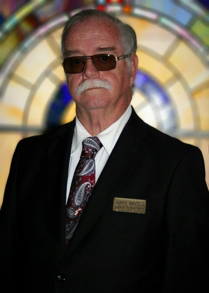 Man in black suit and sunglasses standing before a colorful circular stained-glass backdrop