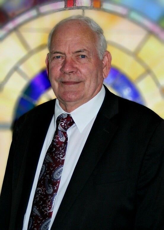Portrait of an older man in a black suit and patterned tie before a colorful stained-glass background