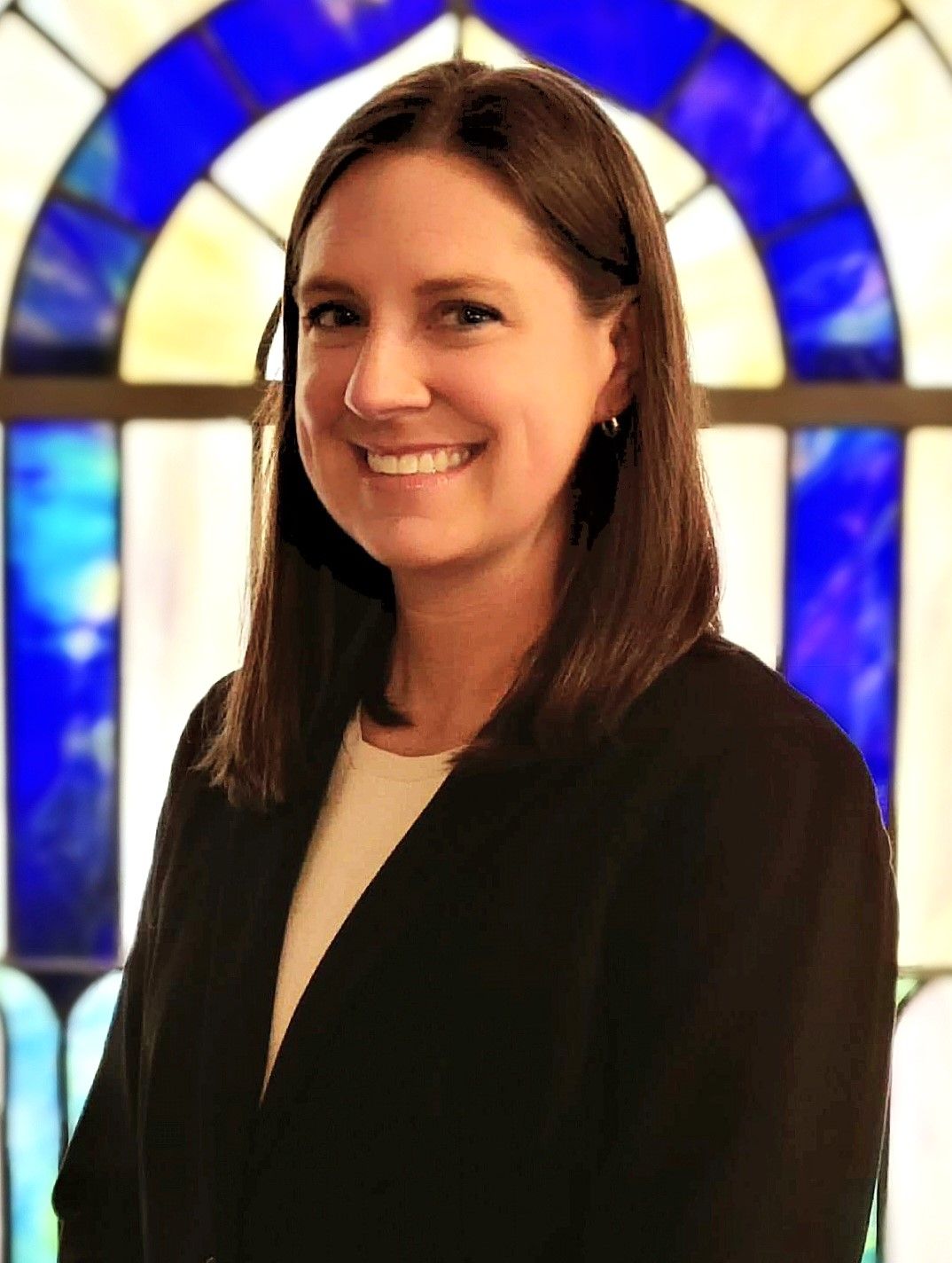 Smiling woman in a black blazer standing before a blue and amber stained-glass window