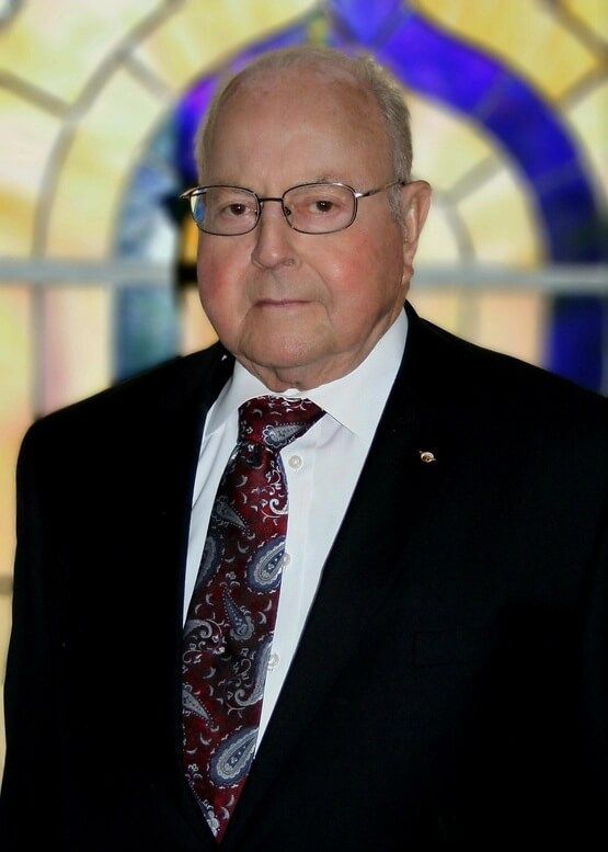 Man in a dark suit, patterned tie, and glasses standing before a stained-glass window
