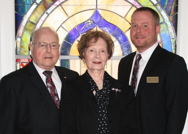 Three people posing in front of a stained-glass window, wearing formal attire.