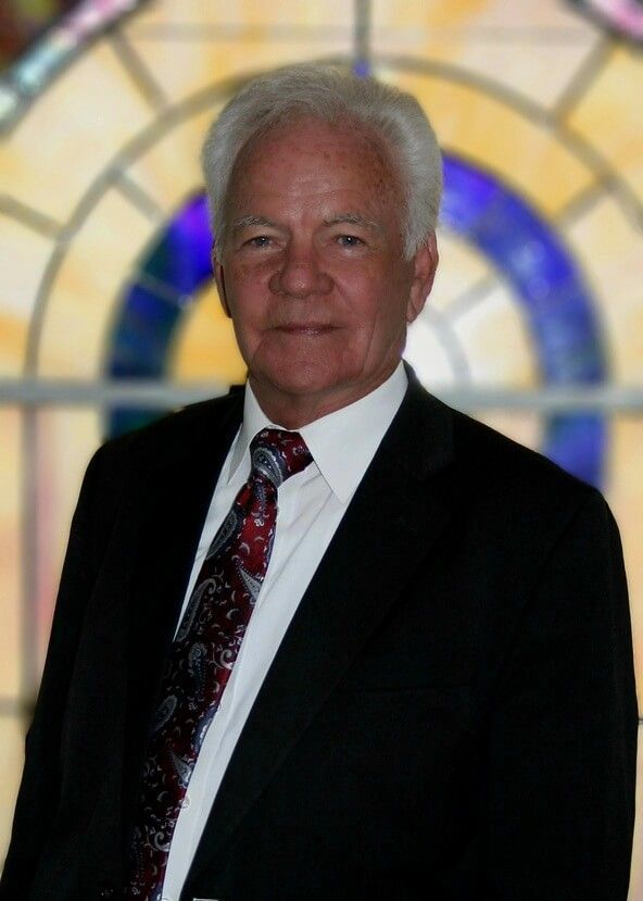 Man in a dark suit and patterned tie standing before a stained-glass window