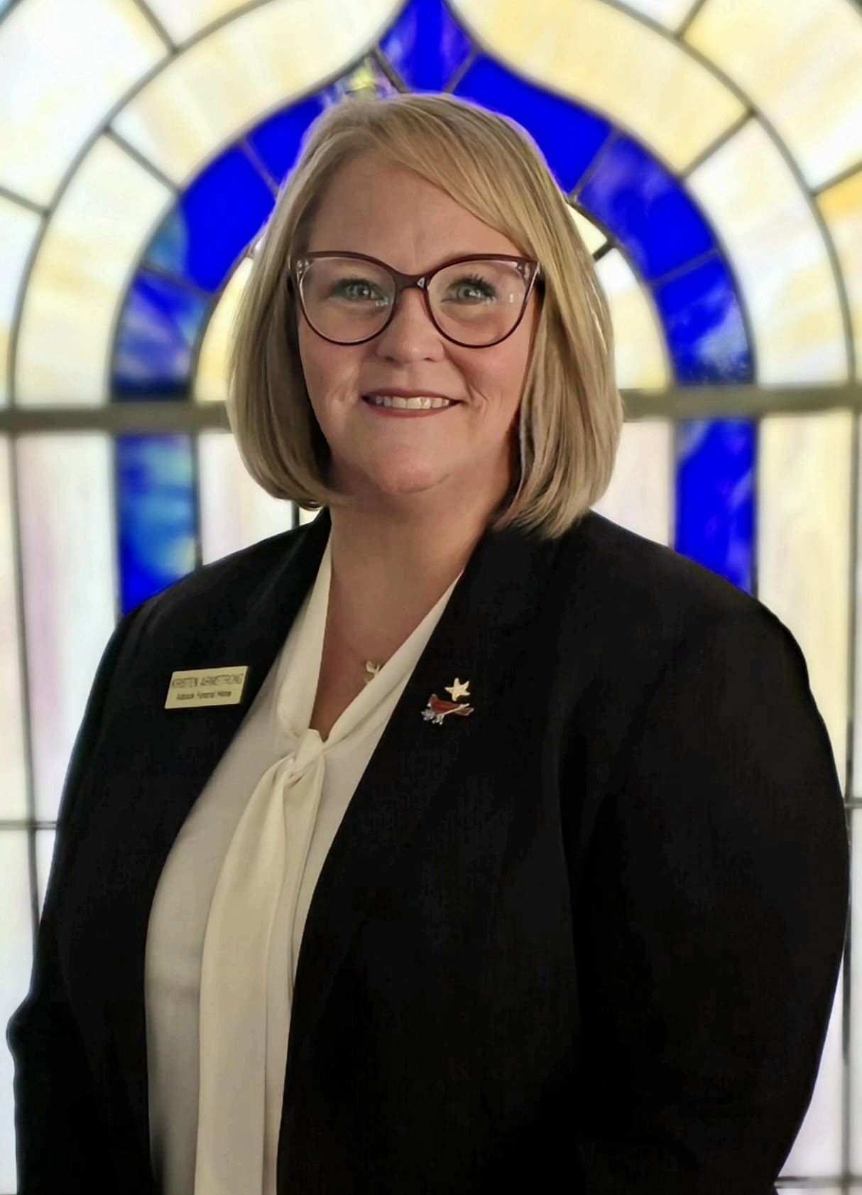Woman in black blazer and glasses standing before a stained-glass window, smiling.