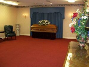 Funeral viewing room with casket, blue drapes, flowers, and red carpet