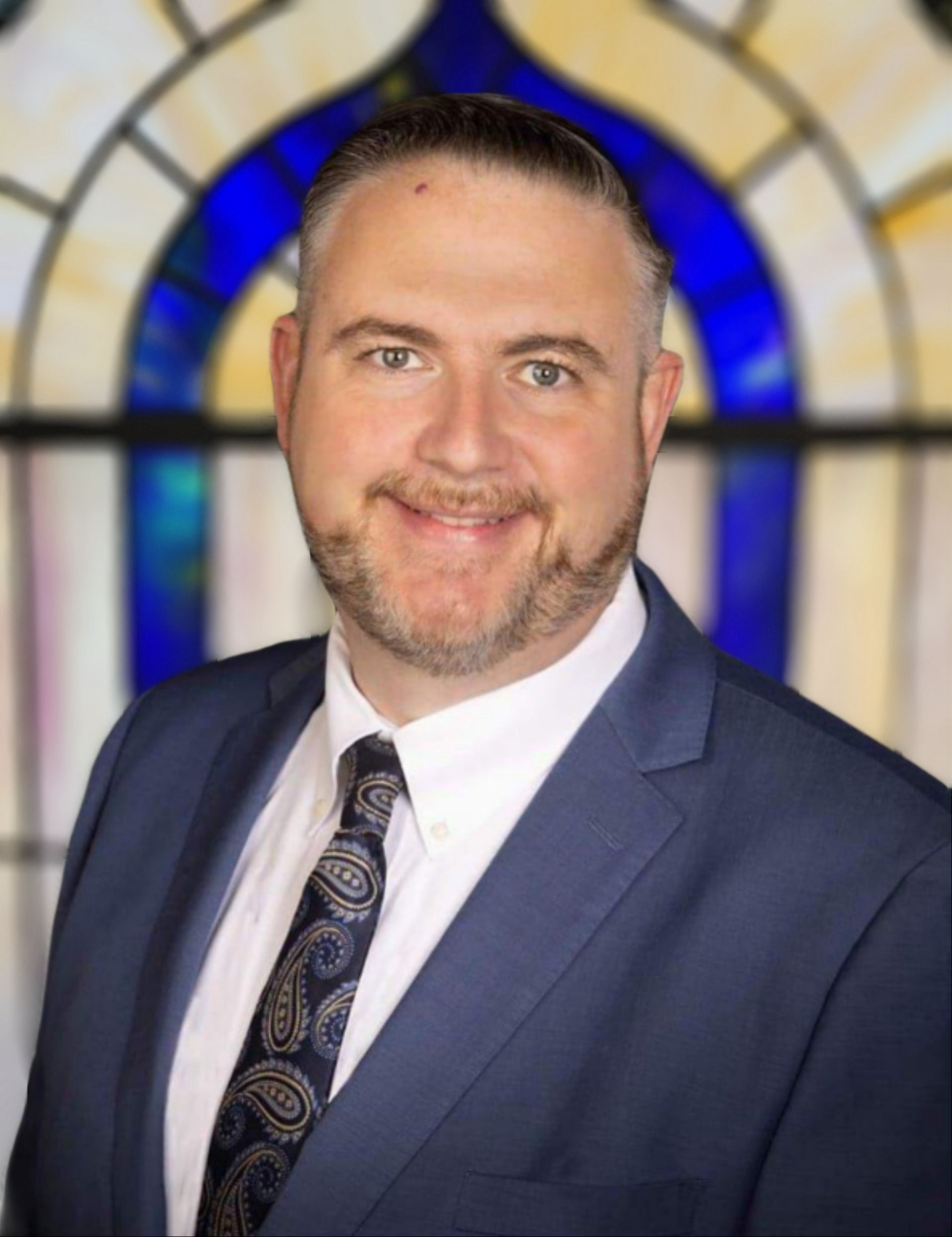 Man in a navy suit and patterned tie smiling in front of a stained-glass window