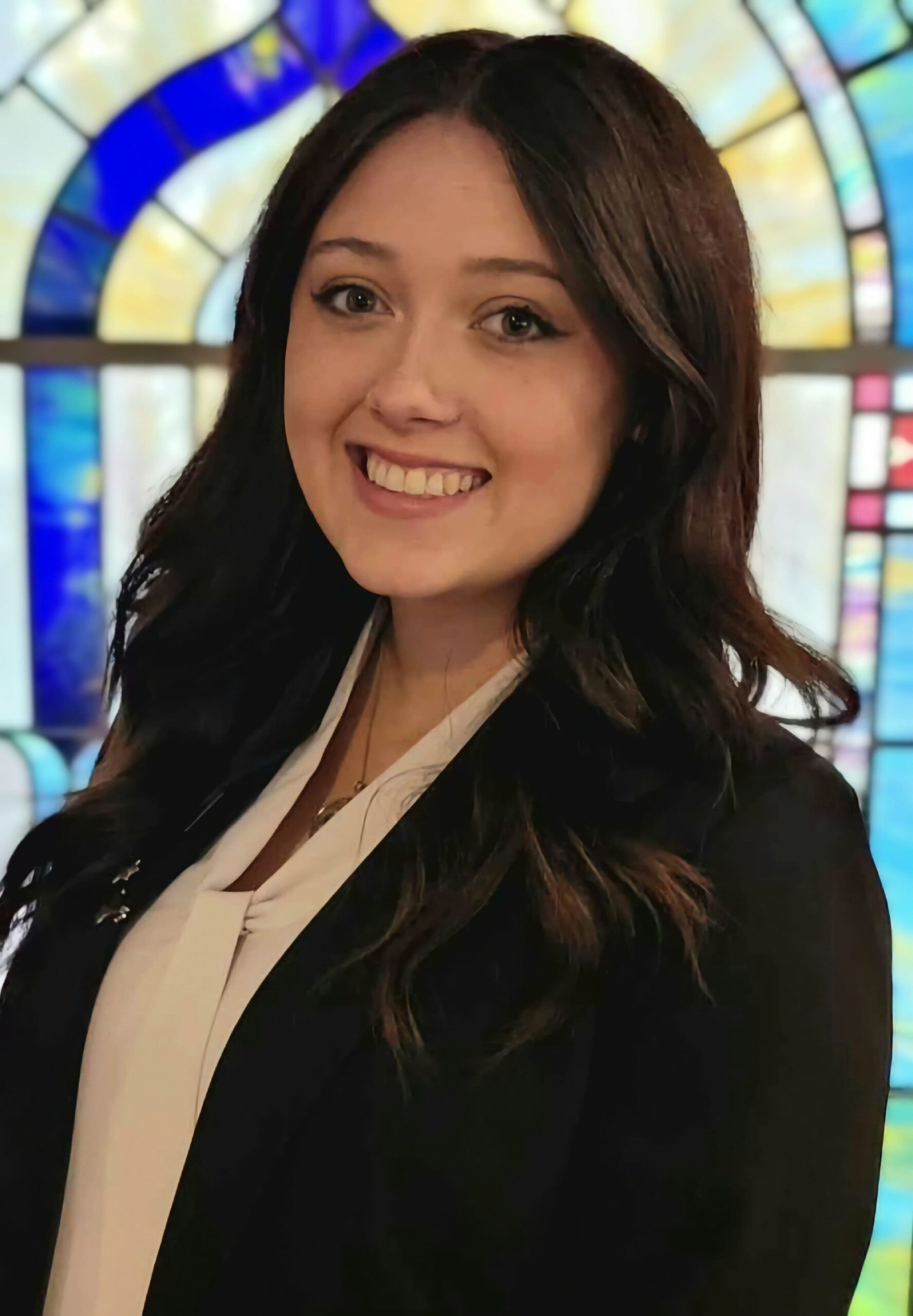 Smiling woman in a black blazer stands before a colorful stained-glass window.