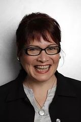 Smiling person with glasses in a black blazer and necklace against a light background