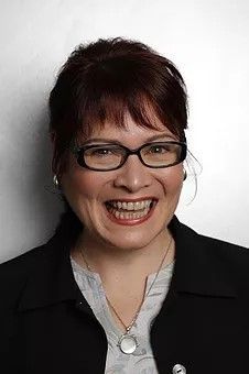 Smiling woman with glasses in a black blazer and silver necklace against a light background