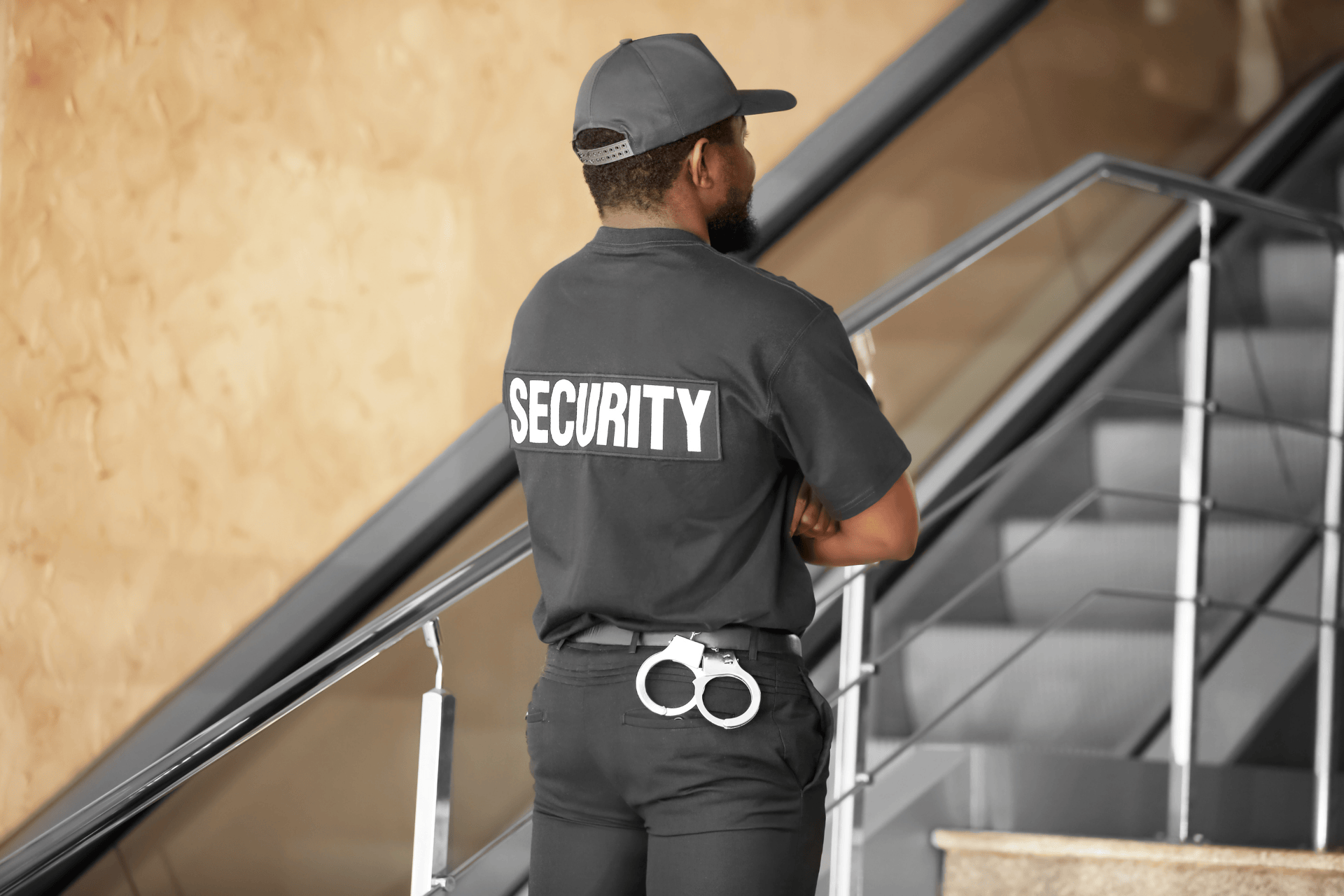 Security guard standing near an indoor staircase, observing surroundings while on duty, wearing a uniform with “SECURITY” on the back and carrying handcuffs.