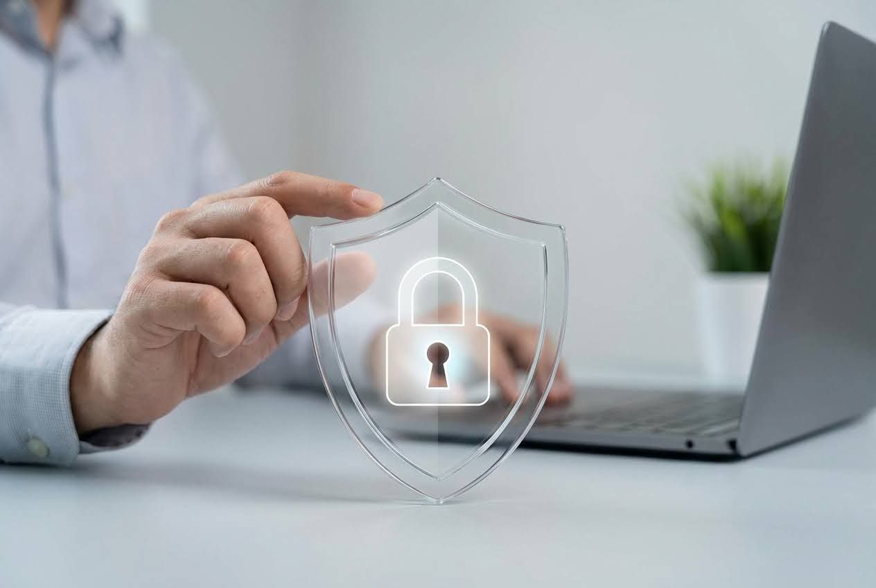  Hand holding a clear shield with a glowing white padlock icon, with a laptop and plant in the background.