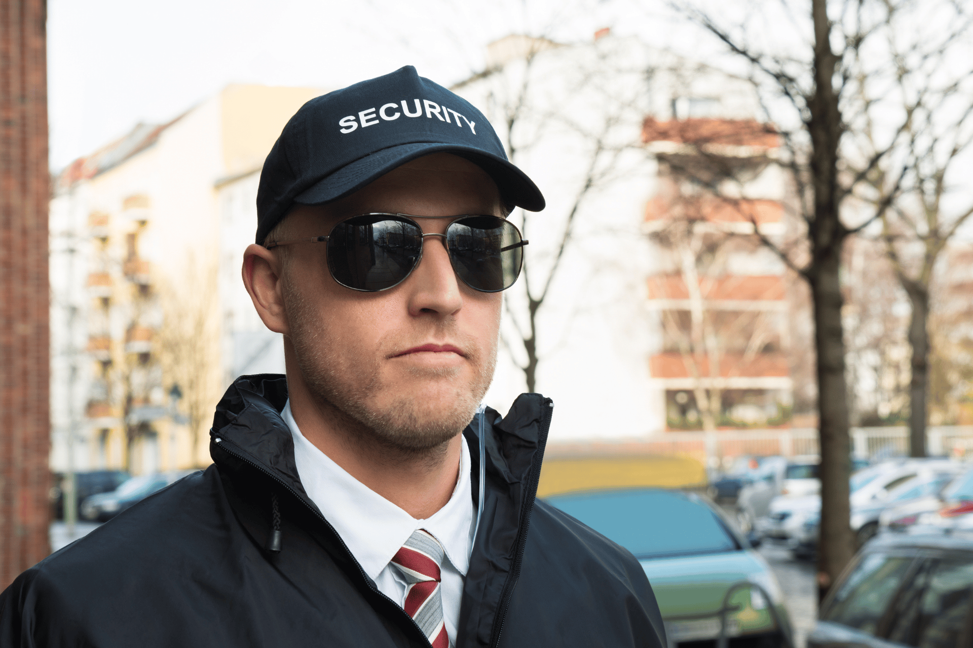 Man in black cap and sunglasses standing outdoors on a city street, wearing a dark coat and striped tie.