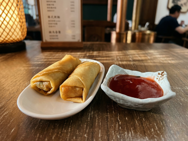 Two golden-brown spring rolls on an oval plate next to a small bowl of red dipping sauce on a wooden restaurant table.