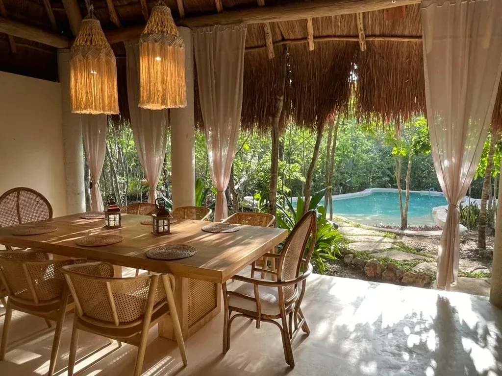 Comedor con mesa y sillas de madera, abierto a una piscina y exuberante vegetación, bajo un techo de paja.