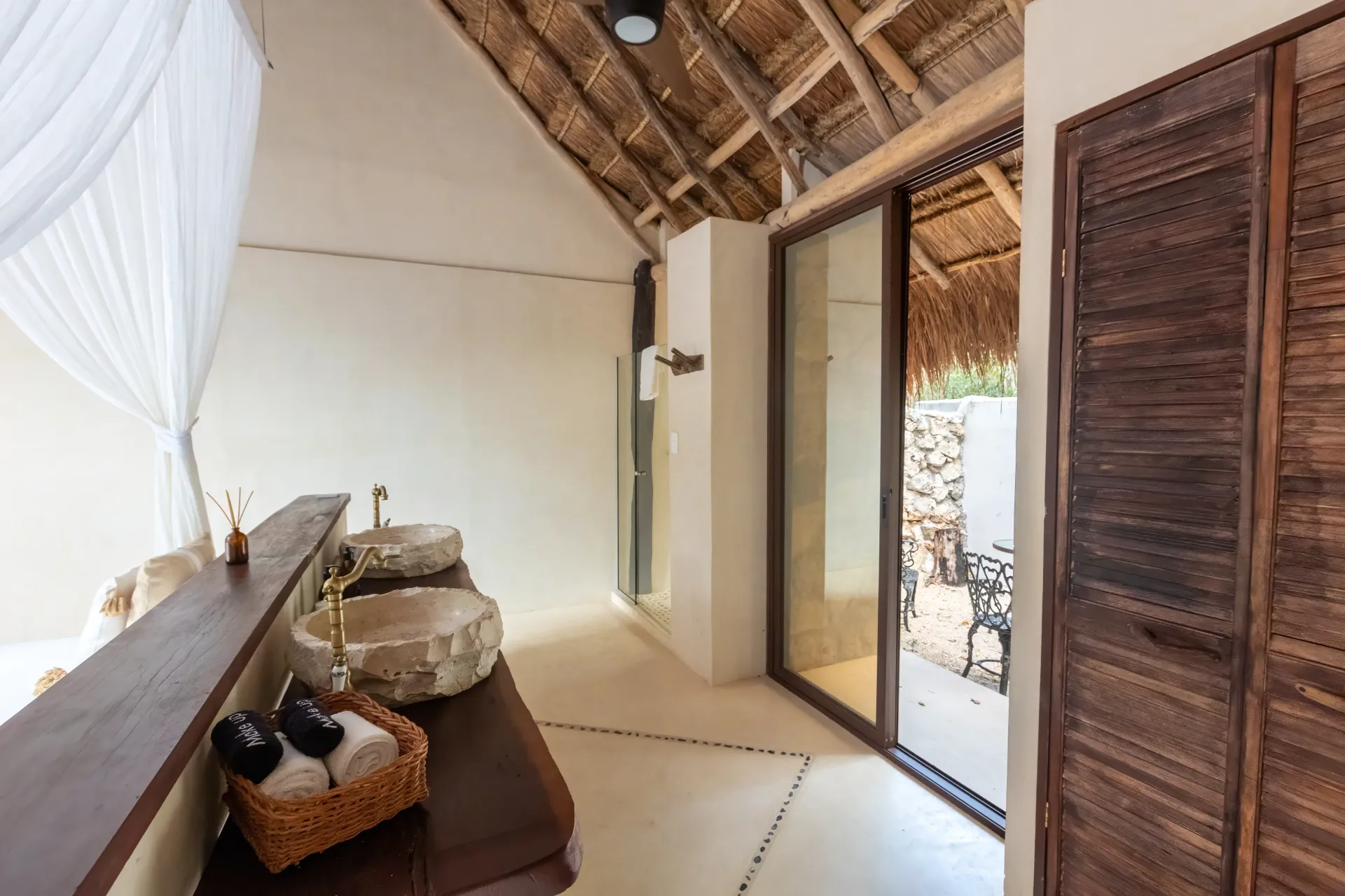 Baño con lavabos de piedra, techo de paja y puerta corrediza de vidrio al área exterior.