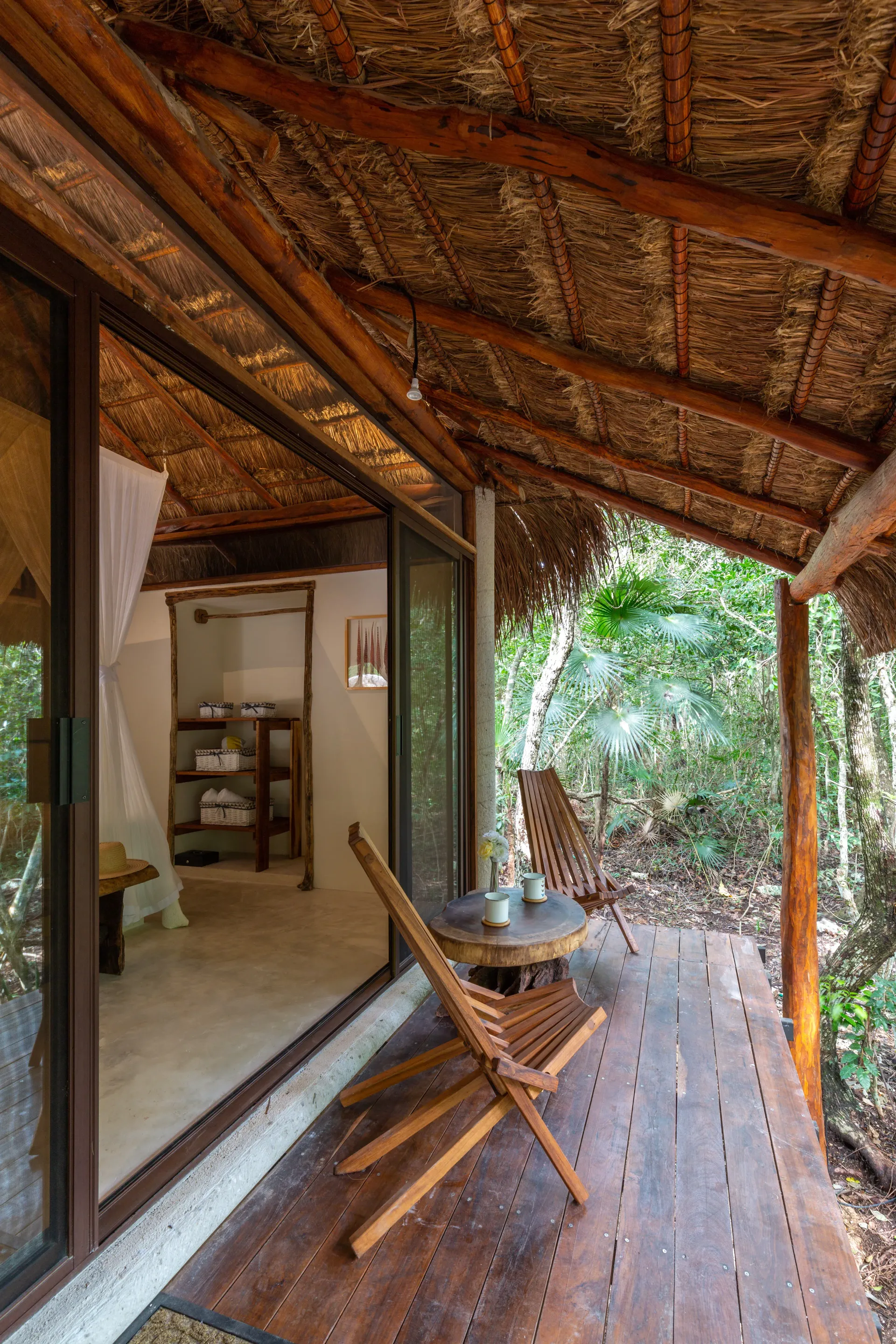 Cabaña de madera con techo de paja, con vistas al bosque. Hay una silla en la terraza.