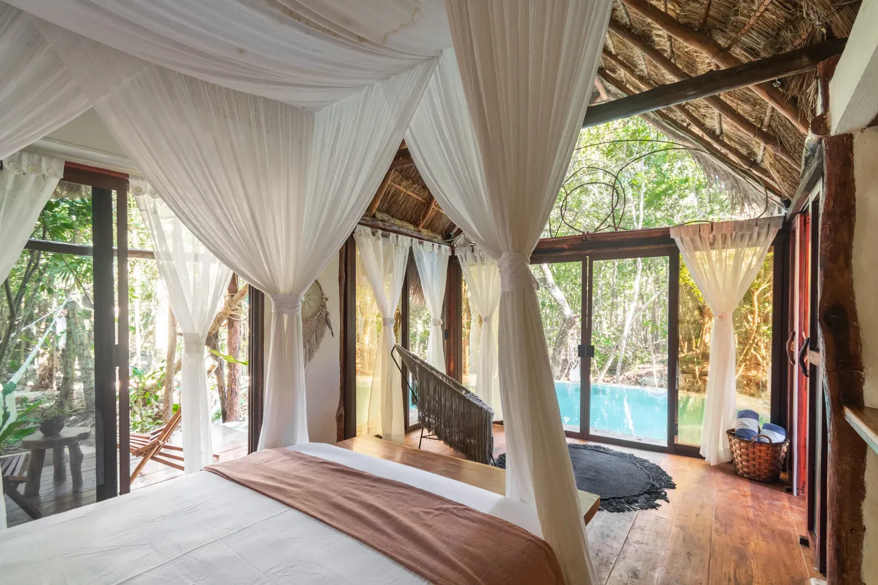 Dormitorio con cama con dosel, grandes ventanales con vistas a la piscina y zona verde.