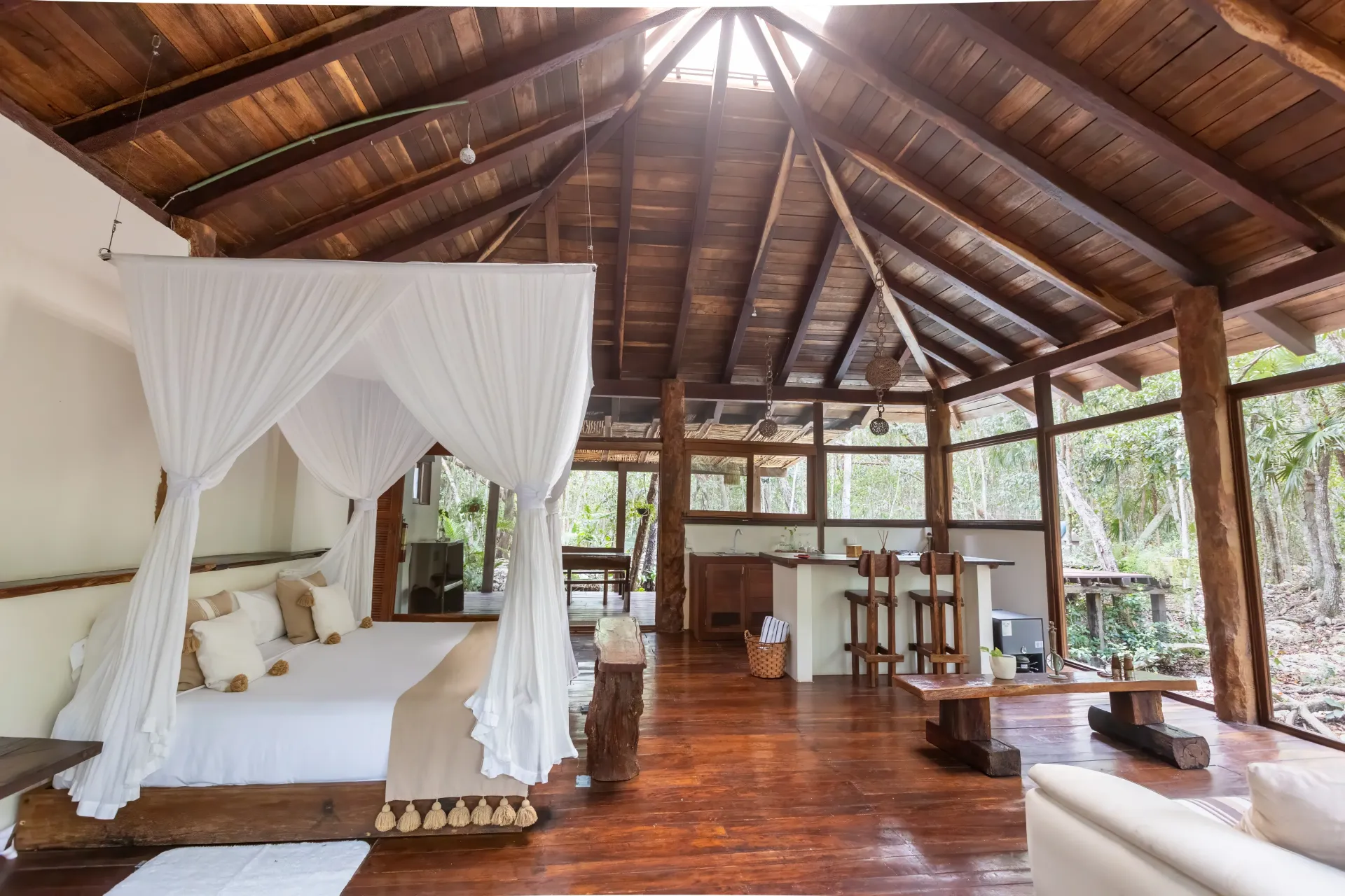 Interior de cabaña de madera con cama, dosel y cocina al aire libre, con vista a una zona boscosa.