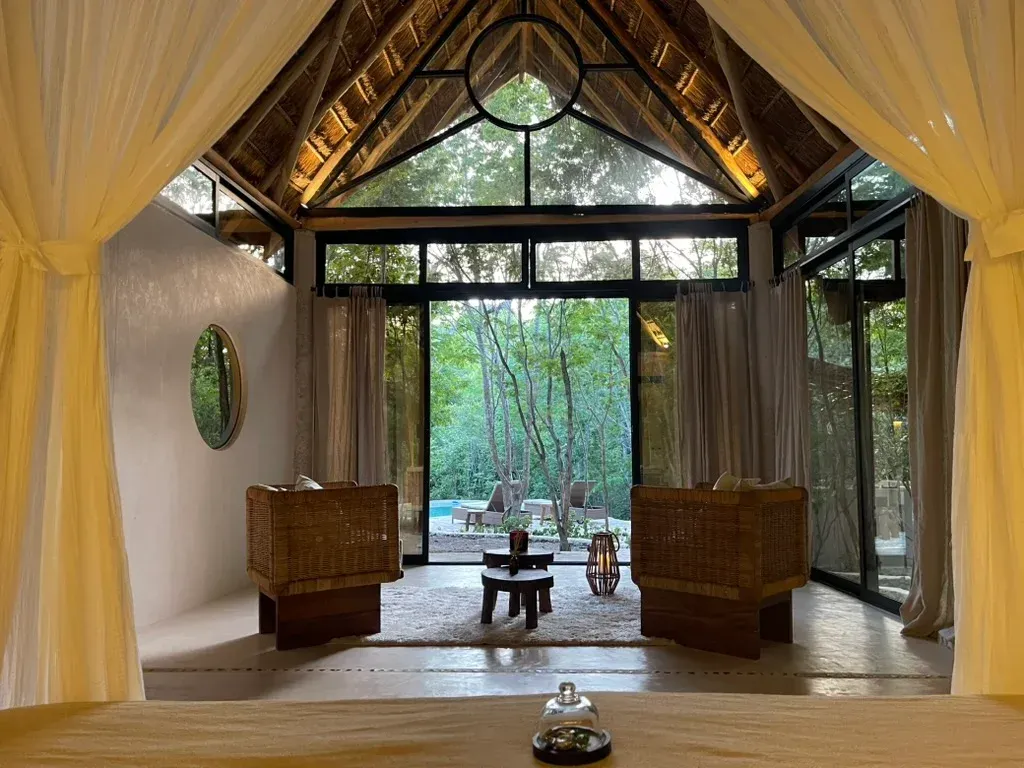 Interior de sala de spa con sillas tejidas, puertas abiertas a la vegetación y cortinas drapeadas.