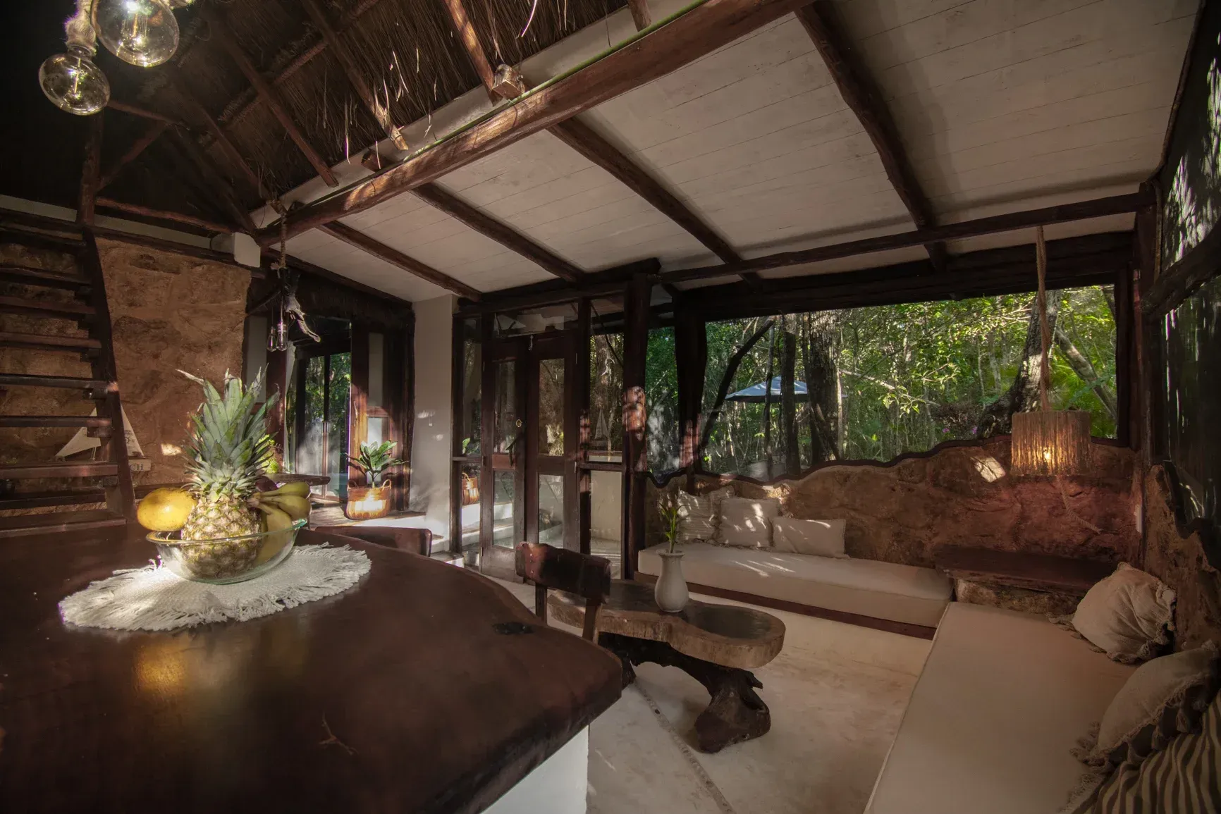 Interior de una cabaña rústica con amplios ventanales con vistas al bosque. Muebles marrones, cojines blancos y luz natural.