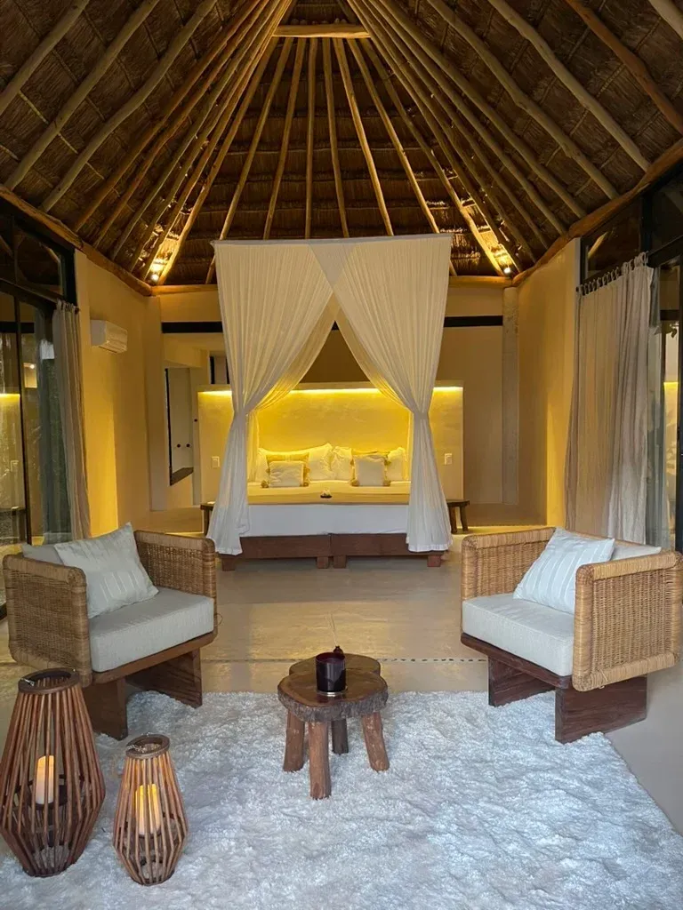 Acogedor salón al aire libre con sillas tejidas, una pequeña mesa de madera y una cama con dosel, bajo un techo de paja.