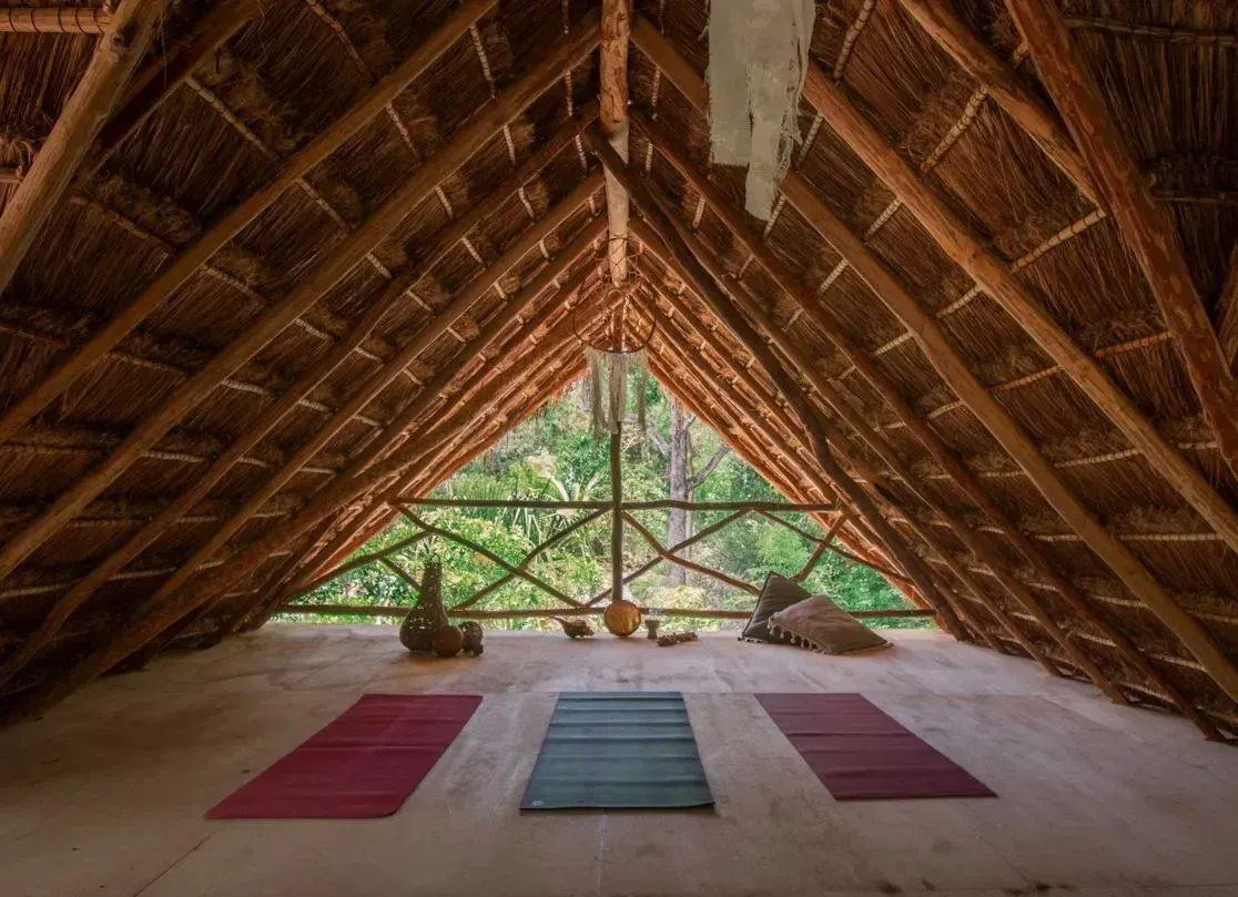 Estudio de yoga con techo de paja y vistas a un exuberante paisaje. Tres esterillas en el suelo.