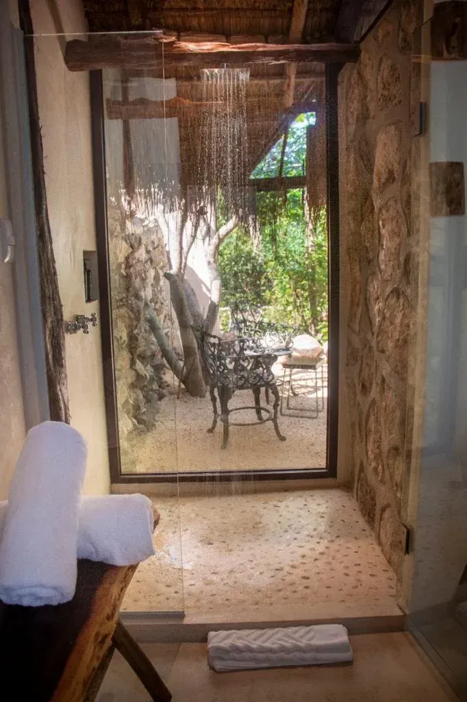 Ducha exterior con paredes de piedra y vistas a un jardín, con toallas blancas sobre un banco.