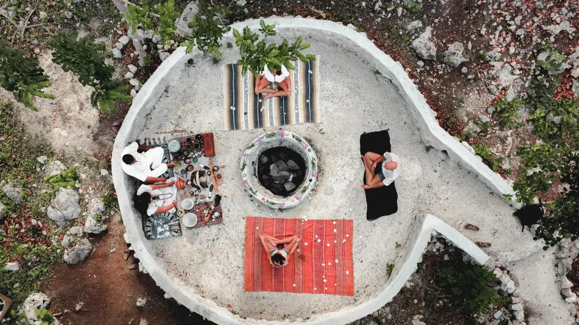 Vista aérea de un espacio blanco al aire libre con gente descansando sobre alfombras alrededor de una fogata central.