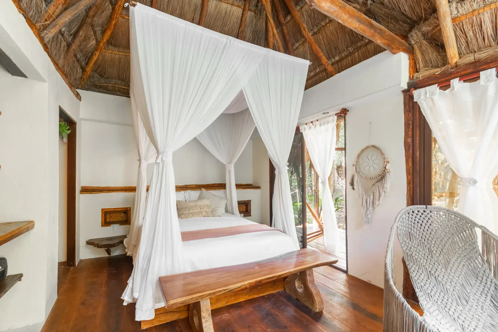 Dormitorio con cama con dosel, banco de madera y silla tejida, ambiente tropical.
