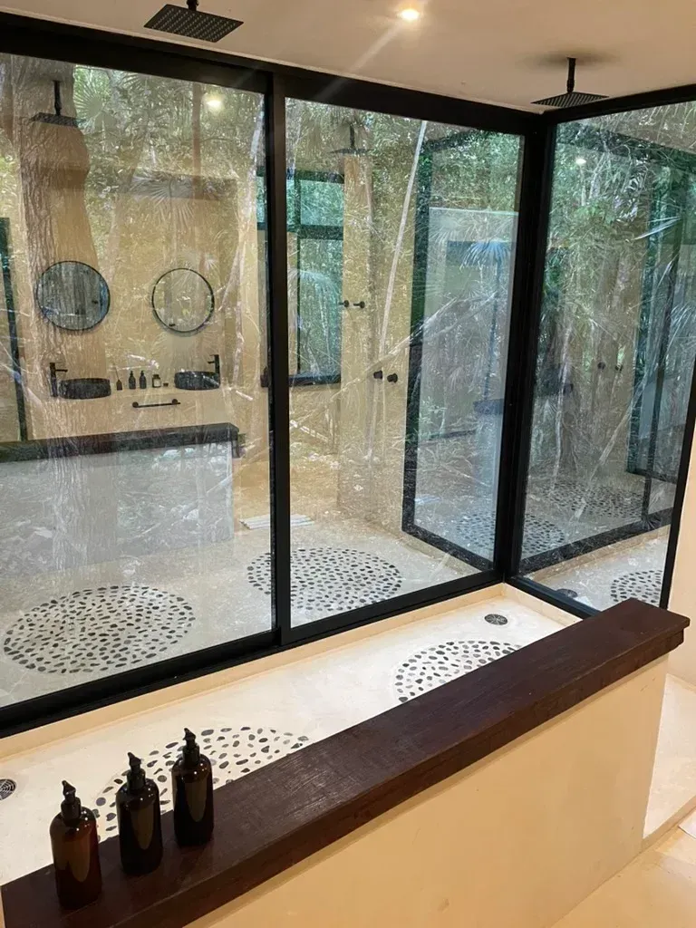 Baño con paredes de cristal, marcos negros, estante de madera oscura con botellas y zona de lavabo.