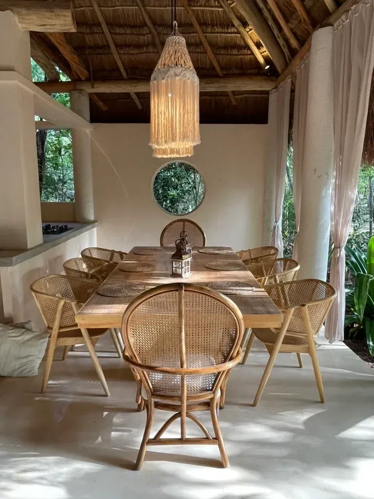Comedor: mesa de madera, sillas tejidas, farol, lámpara colgante y árboles al fondo.