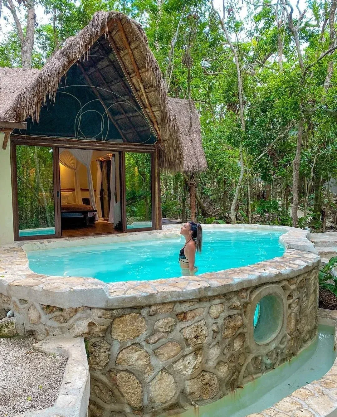 Mujer en una piscina frente a una cabaña. La piscina está rodeada de piedra. Frondosos árboles rodean la zona.
