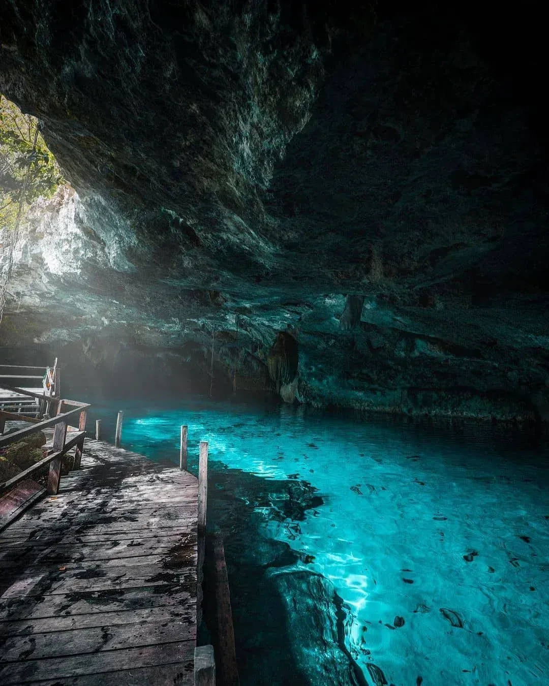 Pasarela de madera junto a las aguas turquesas de una cueva; la luz del sol entra a raudales.