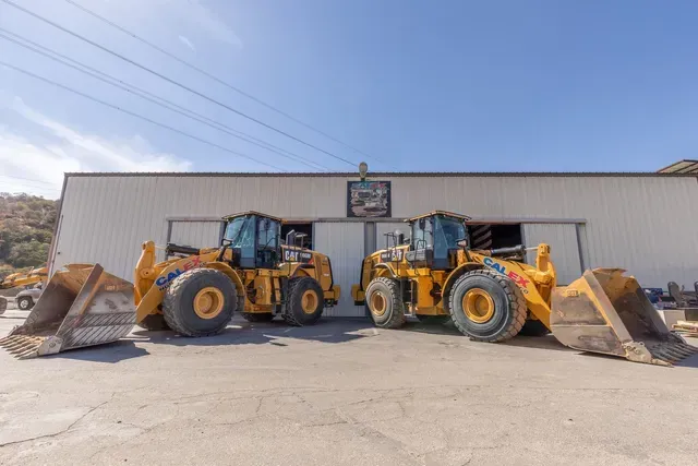Two yellow front loaders parked outside a warehouse, with buckets raised and wide tires on a sunny day.