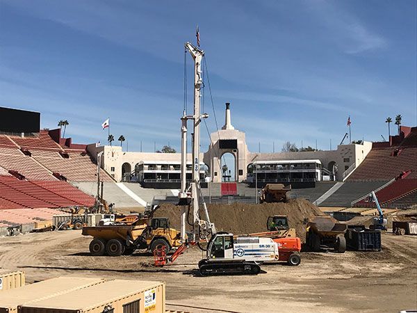 Construction equipment on a stadium field with a tall drilling rig and empty red stands in the background