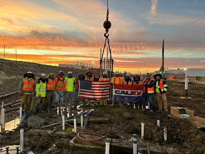 Construction crew in safety vests holding a U.S. flag and CALI banner at a sunset drilling site